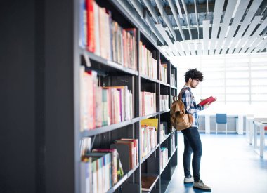 Erkek öğrenci kütüphanede, kitap arıyor, final sınavına hazırlanıyor. Üniversite kütüphanesindeki raflardaki kitaplar, ders kitapları, günlükler.