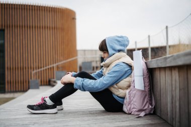 Sad girl alone, sitting in front of school building, waiting for parents. Young schoolgirl with no friends, feeling lonely. Mental health problems. clipart