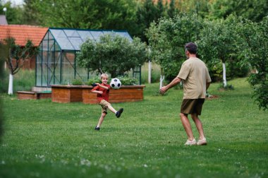 Babamın küçük oğluyla eğlenmesi, futbol oynaması, koşması ve top oynaması. Evlerinin önündeki çimenlikte oynuyorlar. Babalık ve Babalar Günü konsepti.