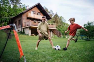 Babamın küçük oğluyla eğlenmesi, futbol oynaması, koşması ve top oynaması. Evlerinin önündeki çimenlikte oynuyorlar. Babalık ve Babalar Günü konsepti.