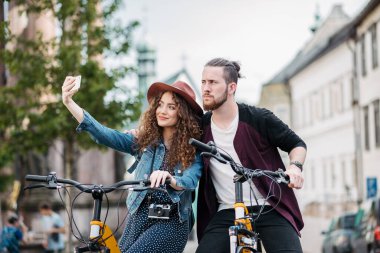 Gezgin çift selfie çekiyor, şehirde elektrikli scooterların yanında dikilip geziyorlar. Yaz tatillerinde bisiklet kullanan genç turistler..