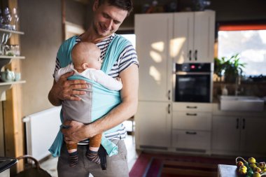 Babası kucağında küçük bir bebek, onu mavi bebek taşıyıcısı ile evin içinde taşıyor. Koşulsuz baba sevgisi, Babalar Günü konsepti.