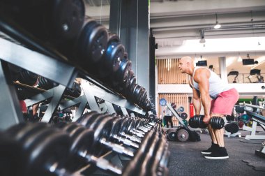 Tek kollu dambıl sırasını eğik pozisyonda yapan adama yaklaş. Güç egzersizi. Fiziksel ve zihinsel sağlık için rutin çalışma.