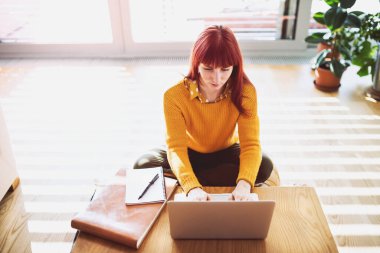 Kızıl saçlı kadın ofisten çalışırken dizüstü bilgisayarda yazı yazıyor. Şehirdeki dairesinden uzaktan çalışan iş kadını..