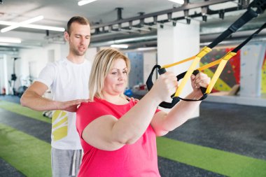 Kilolu bir kadın spor salonunda egzersiz yapıyor, uzaklaştırma eğitimi alıyor, tamamen direnç egzersizi yapıyor. Kişisel antrenör onu çalıştırıyor ve ona yardım ediyor.