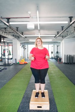 Kilolu bir kadın spor salonunda, halterlerle egzersiz yapıyor.