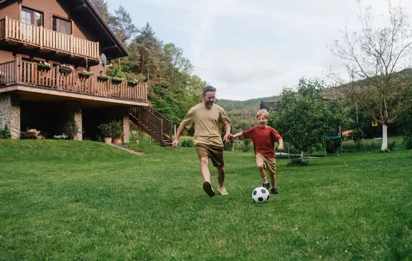 Babamın küçük oğluyla eğlenmesi, futbol oynaması, koşması ve top oynaması. Evlerinin önündeki çimenlikte oynuyorlar. Babalık ve Babalar Günü konsepti.