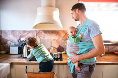 Baba kucağında küçük bir bebek askısı tutuyor. Büyük kardeş tencerede lapa karıştırıyor. Koşulsuz ebeveyn sevgisi ve aile anı.