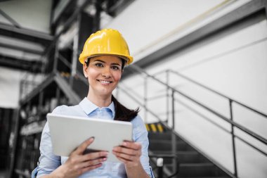 Modern endüstriyel fabrikada elinde tablet olan kadın proje yöneticisinin portresi. Robot, robotik kollar ve otomasyon ile üretim tesisi.