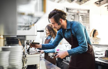 Kahve dükkanında çalışan iki genç barmen tezgahta durup makinede kahve hazırlıyorlar. Üniversite öğrencileri bir kafede yarı zamanlı çalışıyorlar..