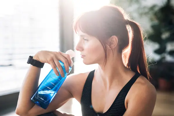 stock image Beautiful redhead woman drinking water, resting after home fitness routine. Home workout on gym mt in living room, sporty woman wearing sportswear clothes.