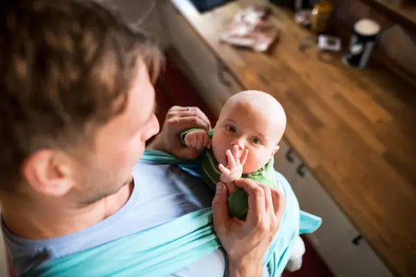 stock image Father holding small baby, carrying him around house in blue baby carrier. Unconditional paternal love, fathers Day concept.