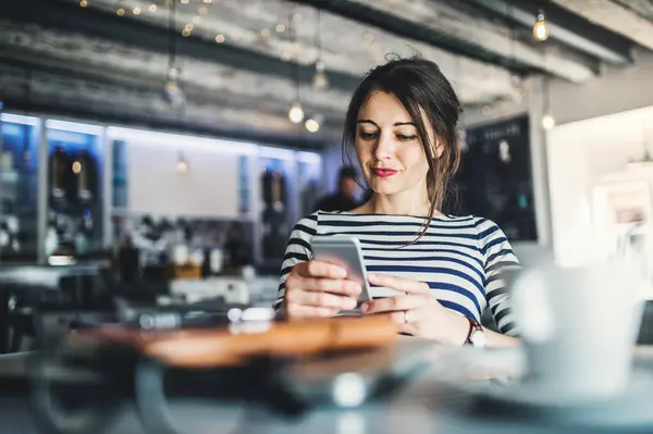 stock image Woman working remotely from coffee shop. Cappucino and laptop on table. Female freelancer scrolling on smartphone in cafe.