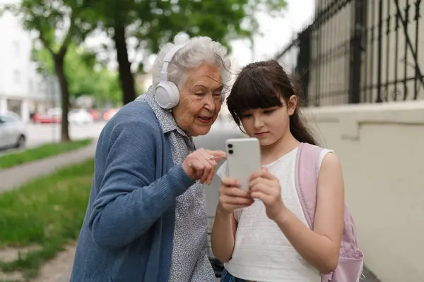 stock image Grandma picking up young girl from school at afternoon. Granddaughter showing funny video on smartphone to senior grandmother with headphones.