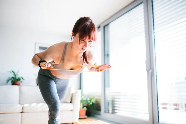 stock image Beautiful redhead womans home fitness routine with resistance band. Home workout on gym mat in living room, sporty woman wearing sportswear clothes.