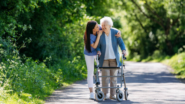 Caregiver pushing senior woman in wheelchair. Nurse and elderly woman enjoying a warm day in nursing home, public park.