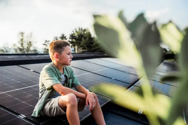 stock image Cute boy sitting on roof with solar panels, looking at surrounding nature. Sustainable future for next generation concept.