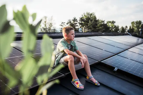 stock image Cute boy sitting on roof with solar panels, looking at surrounding nature. Sustainable future for next generation concept.