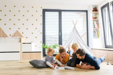 Young nuclear family playing with toys in a living room. Parents and children lying on floor, looking at childrens story, picture book, spending weekend day indoors. clipart