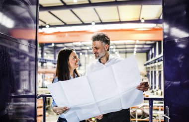 Two project managers standing in modern industrial factory, looking at blueprints. Manufacturing facility with robotics, robotic arms and automation. clipart