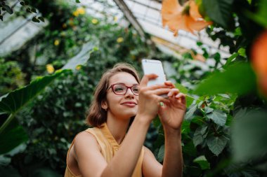 Botanik bahçesindeki bir kadın, yemyeşil yaprakların ortasında, elinde akıllı telefon, çiçeğin fotoğrafını çekiyor..