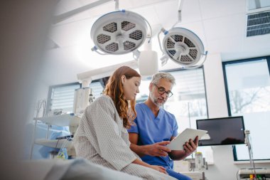 Doktor test sonuçlarını genç bir hastayla tartışıyor, acil serviste tablette verileri gösteriyor..