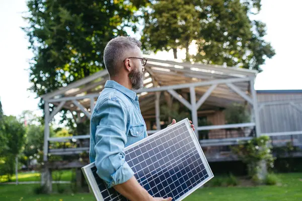 stock image Man carrying solar panel in arms, walking across garden. Sustainable lifestyle and green household, home.