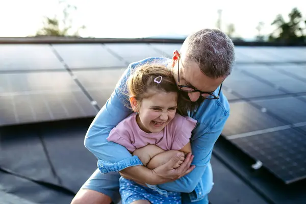 stock image Father with girl on roof with solar panels, hugging. Rooftop solar or photovoltaic system. Sustainable future for next generation concept.