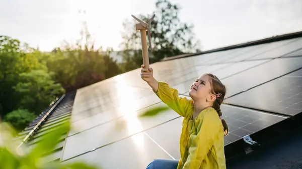 stock image Cute girl on roof with solar panels, holding model of wind turbine. Rooftop solar or photovoltaic system. Sustainable future for next generation concept.