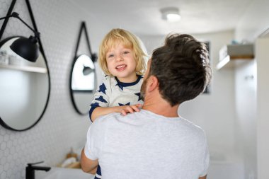 Baba oğlunu kollarında tutuyor, banyoda duruyor. Anaokuluna gitmeden önce genç bir çocuk için sabah rutini.