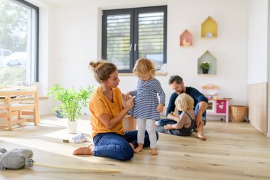 Genç bir nükleer aile oturma odasında oyuncaklarla oynuyor. Ebeveynler ve çocuklar koşuyor, eğleniyorlar, hafta sonunu evde geçiriyorlar..