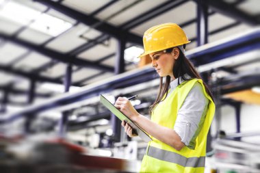 Female warehouse manager woman in reflective safety clothes controlling goods in the warehouse, writing in clipboard. clipart