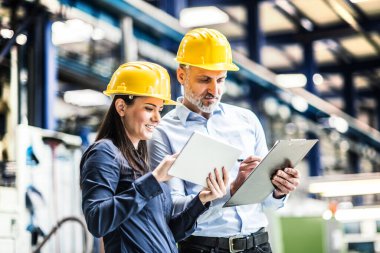 Modern endüstriyel fabrikada duran iki proje yöneticisi, panodaki belgeye bakıyor. Robot, robotik kollar ve otomasyon ile üretim tesisi.