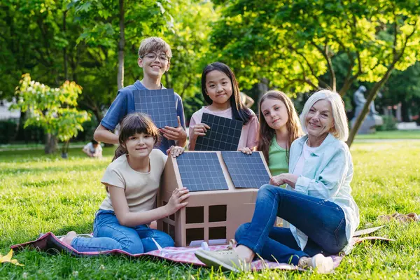 stock image Children learning about renewable solar energy during sustainable education class outdoors, using cardboard model of house wit solar panel on roof.