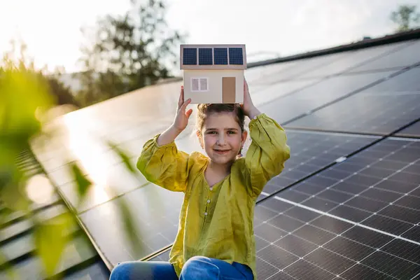 stock image Cute girl holding model of house with solar panels, sitting on rooftop solar or photovoltaic system.