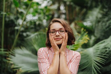 Botanik bahçesinde duran, yemyeşil yaprakların ortasında, yüzünü tutan bir kadın..