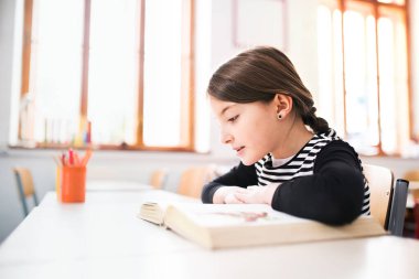 Masada oturmuş, büyük kitap okuyan tatlı bir kızın portresi. İlkokulun sınıfındaki kız öğrenci..