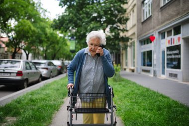 Beautiful elderly woman walking on city street with rollator, going grocery shopping to the store. Making phone call outdoors. clipart