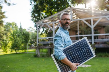 Man carrying solar panel in arms, walking across garden. Sustainable lifestyle and green household, home. clipart