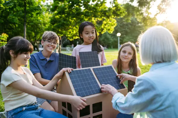 stock image Children learning about renewable solar energy during sustainable education class outdoors, using cardboard model of house wit solar panel on roof.