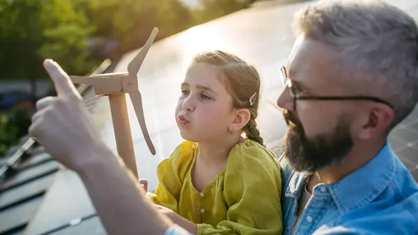 stock image Daughter and father on roof with solar panels, holding model of wind turbine. Rooftop solar or photovoltaic system. Sustainable future for next generation concept.