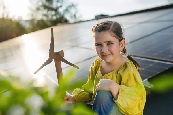 stock image Cute girl on roof with solar panels, holding model of wind turbine. Rooftop solar or photovoltaic system. Sustainable future for next generation concept.