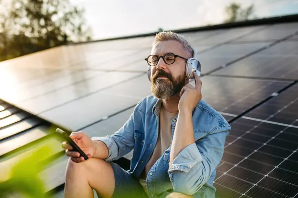 stock image Handsome man sitting on roof full of solar panels, headphones on head, closed eyes.