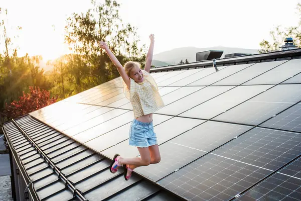 stock image Girl standing on roof with solar panels during sunset, smiling and jumping. Sustainable future for next generation concept.