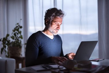 Akşamları ev ofisinden laptopla çalışan yakışıklı bir adam. Ekran ışığı erkeklerin yüzünü aydınlatır. Uzak çalışma.