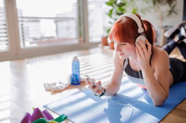 Güzel kızıl saçlı kadın, spor salonundan sonra dinleniyor. müzik dinliyordum. Oturma odasındaki paspasın üzerinde ev çalışması..