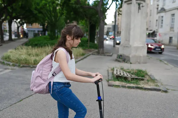 stock image Young girl commuting on scooter to school, riding on the sidewalk with backpack on her back.