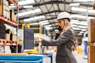 Kadın mühendis, proje yöneticisi bilgisayar başında modern endüstriyel fabrikada duruyor, telefonla konuşuyor. Üretim tesisinde takım yönetimi
