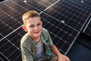 Cute boy sitting on roof with solar panels, looking at camera. Sustainable future for next generation concept. clipart