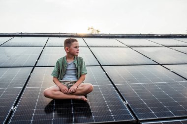 Cute boy sitting on roof with solar panels, looking at surrounding nature. Sustainable future for next generation concept. clipart